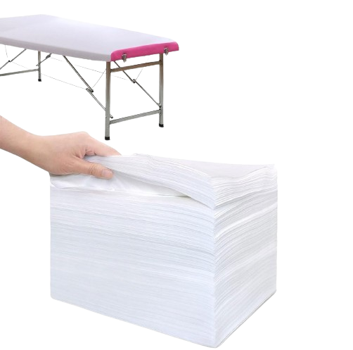 Hand touching a stack of white disposable massage table covers with a portable massage table set up in the background on a plain white background.