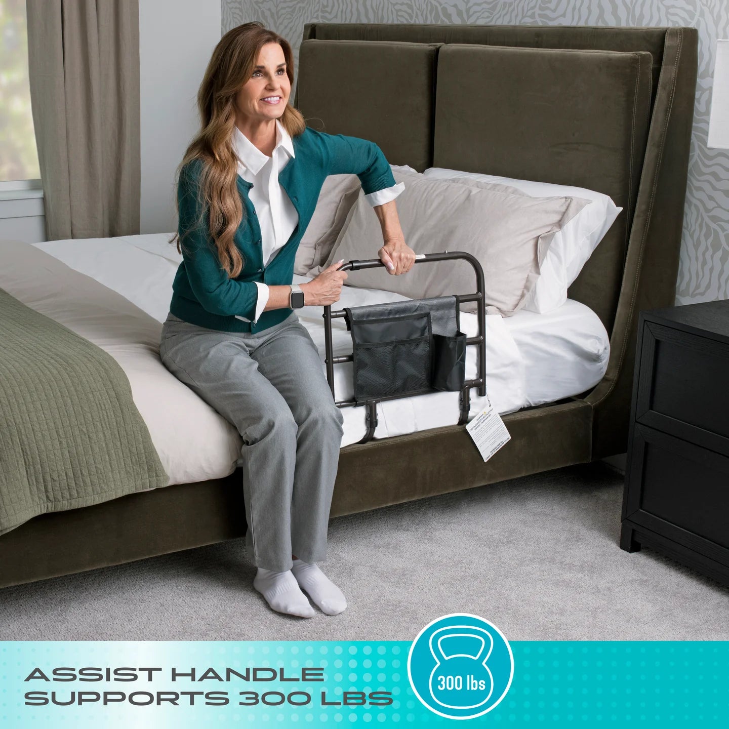 Woman sitting on the edge of a bed using a black Stander Wonder Bed Rail assist handle with organizer pouch to stand up, featuring a teal bottom banner reading ASSIST HANDLE SUPPORTS 300 LBS.
