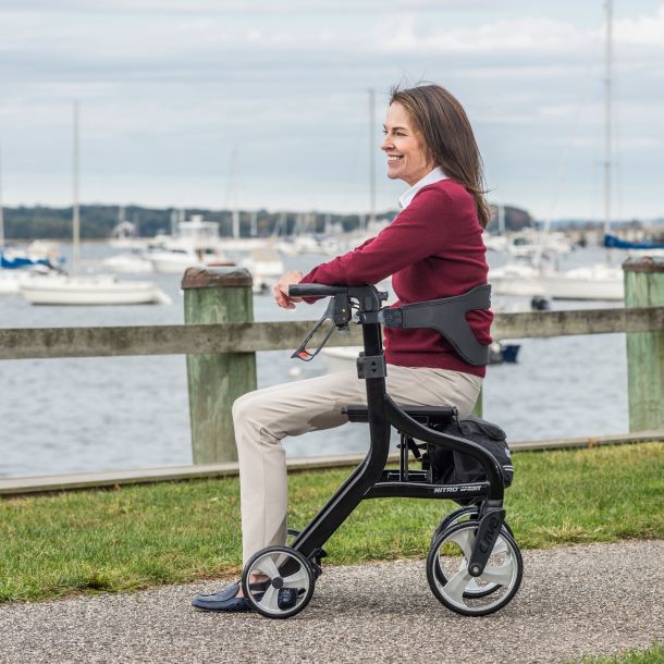 Woman in maroon sweater seated on a black Drive Nitro Sprint rollator walker along a harbor boardwalk with boats in the background.