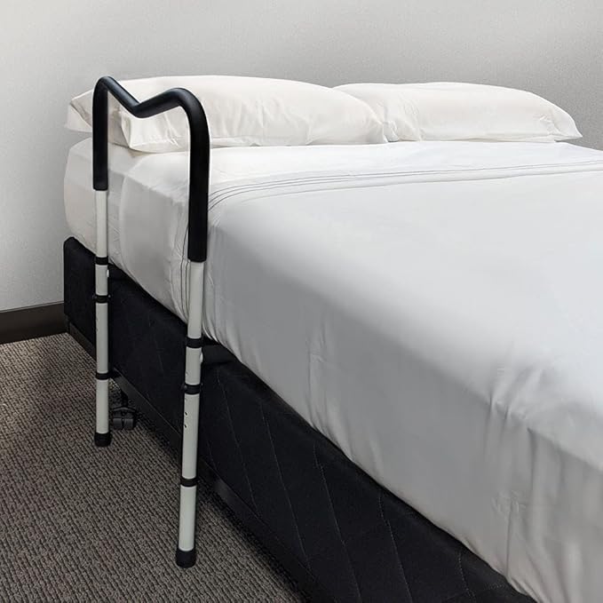 Black and white Avantia adjustable bedside safety rail assist handle installed on the side of a made bed in a bedroom setting.