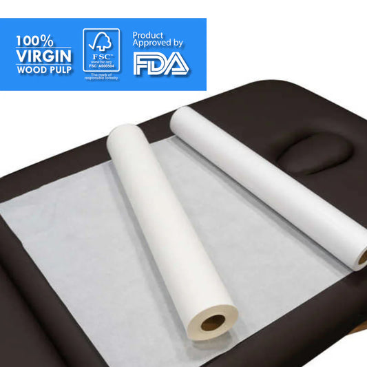 Two rolls of white 100% virgin wood pulp examination table paper unrolled over a dark treatment table, with FSC and FDA logos in the top corner.