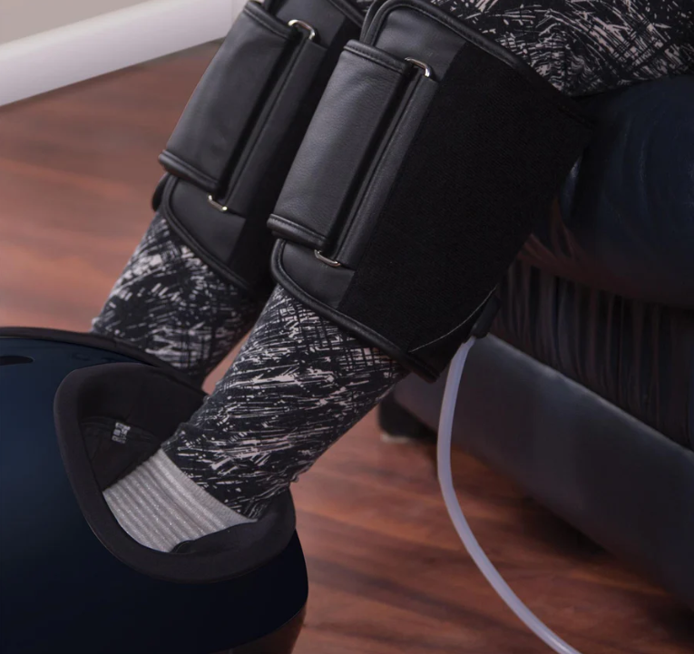 Person sitting down wearing black sequential compression therapy calf wraps attached to a massage device.