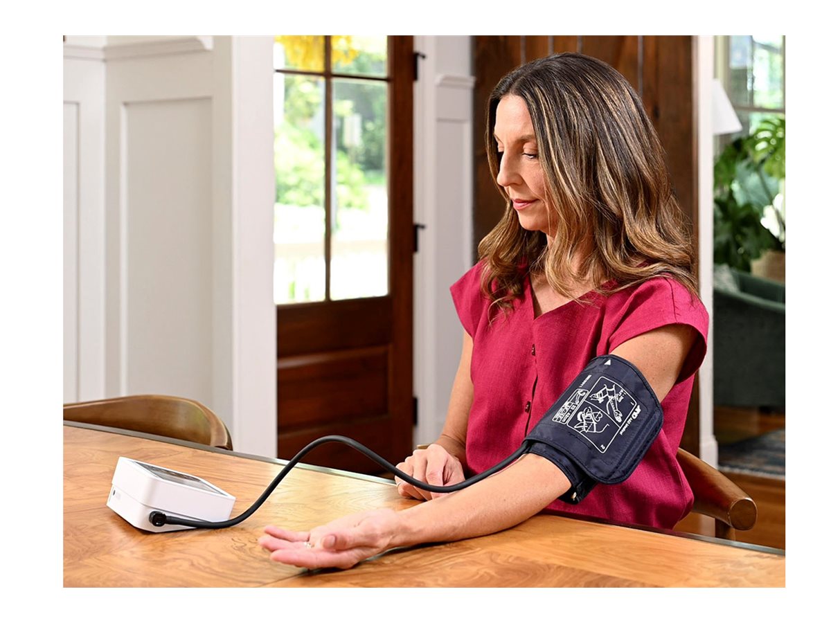 Woman sitting at a wooden table indoors wearing a black cuff and using a digital upper arm blood pressure monitor.