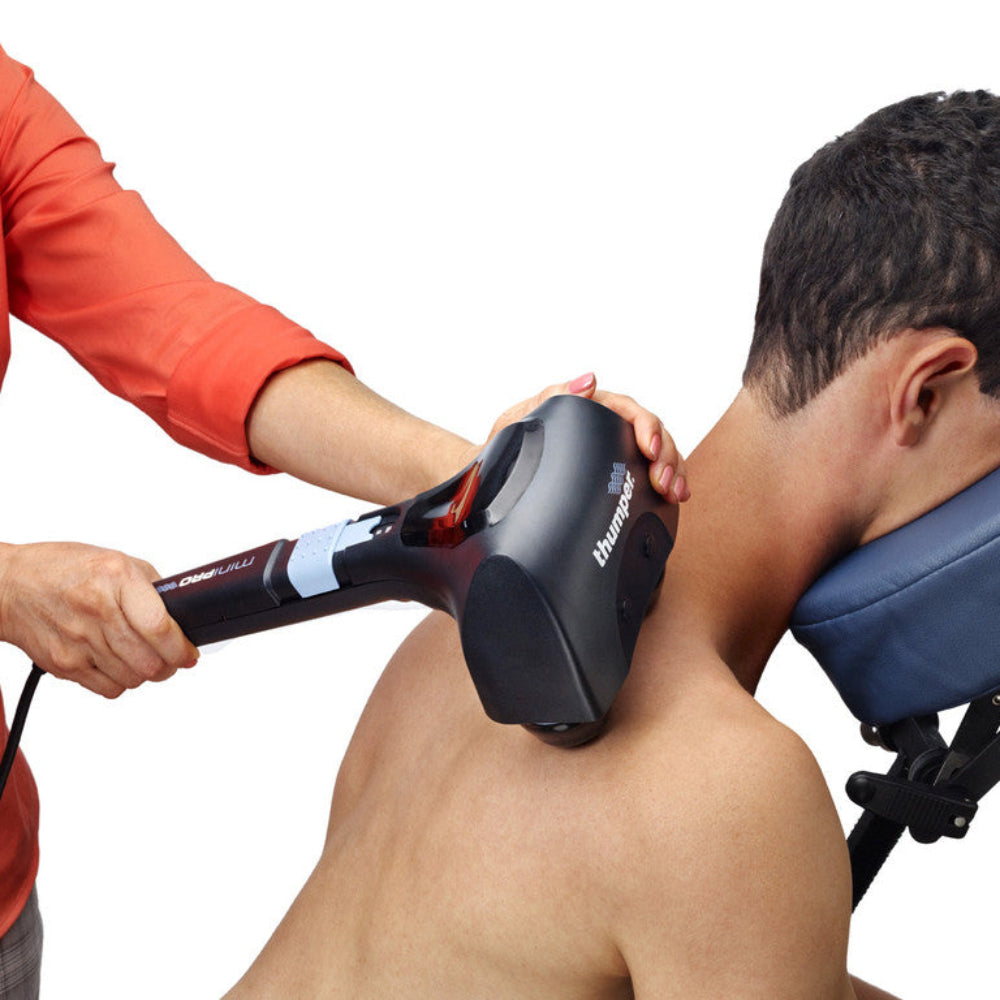 A lifestyle image showing the Thumper Mini Pro being used by a therapist on a patient's upper back and shoulder, demonstrating its effective percussive deep tissue action.